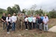 A group of 20 people including 2 men in Army camouflage uniform stand among green vegetation under a grey sky.