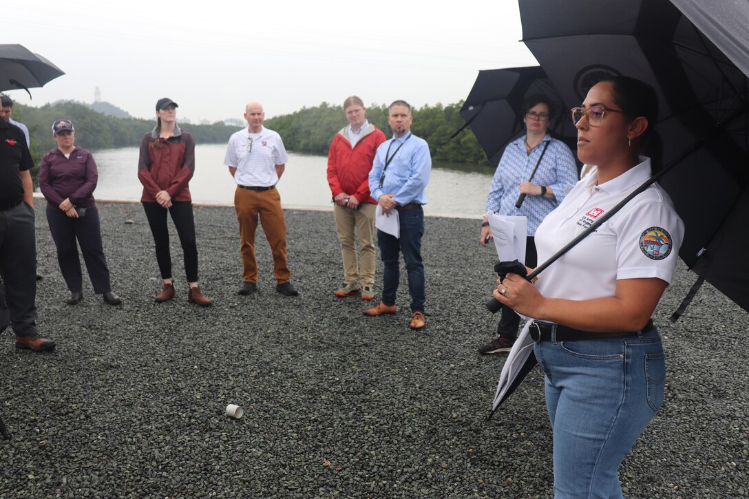 A U.S. Army Corps of Engineers team member in a white polo shirt holding an umbrella addresses a group of 8 people along the bank of a body of water.