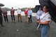 A U.S. Army Corps of Engineers team member in a white polo shirt holding an umbrella addresses a group of 8 people along the bank of a body of water.
