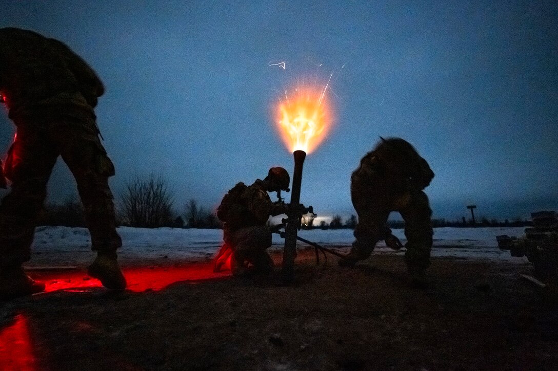 A bright orange flame emits form a mortar system as soldiers reload it during twilight.
