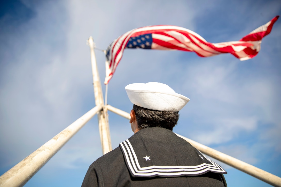 A close-up of the back of a sailor looking up at an American flag blowing in the wind.