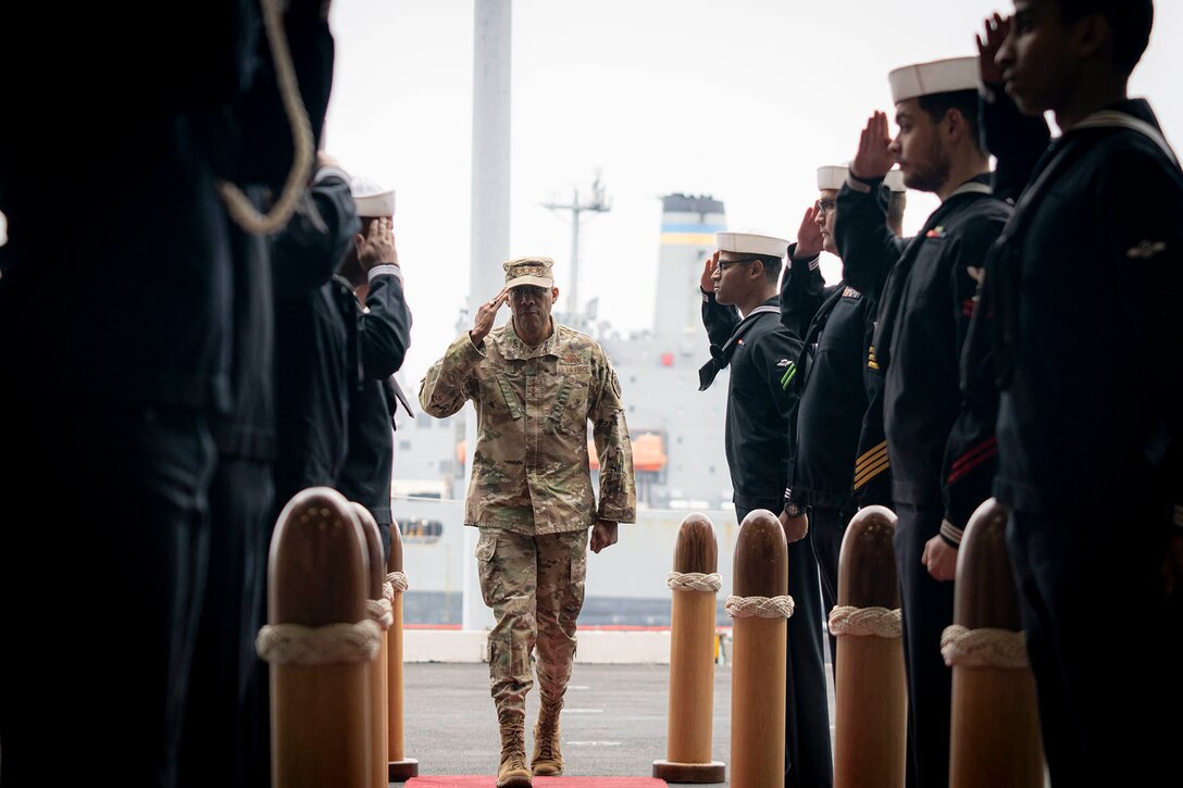 Air Force Gen. CQ Brown, Jr. salutes sailors forming two rows flanking him as he walks aboard a ship.