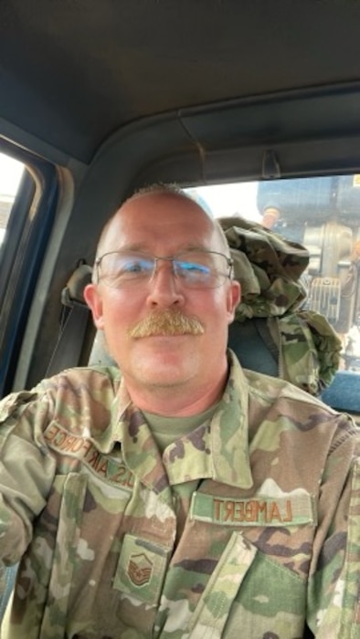 A well-mustachioed man in a camouflage uniform sits in the front seat of a truck.