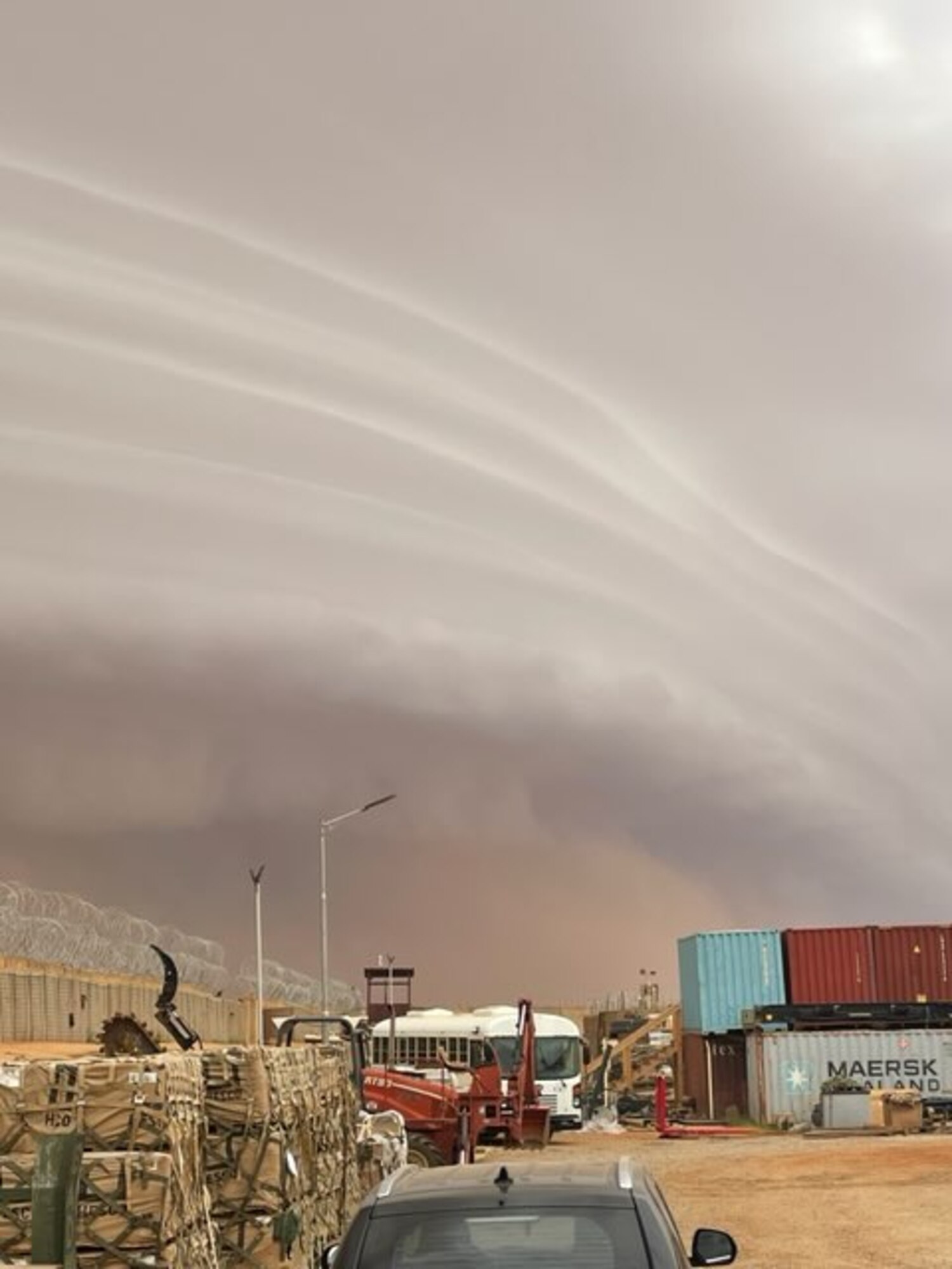 A striated wall of tan clouds looms over a cargo yard surrounded by a razor-wire-topped fence.
