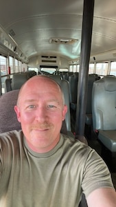 A slightly-mustachioed man in a tan t-shirt sits in the driver's seat of an empty bus.