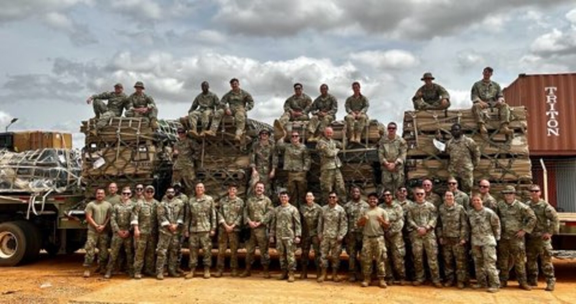 A group of people wearing brown and green camouflage uniforms stand on and in front of a flatbed trailer loaded with four pallets of what appears to be building materials. Some of them have very fine moustaches.