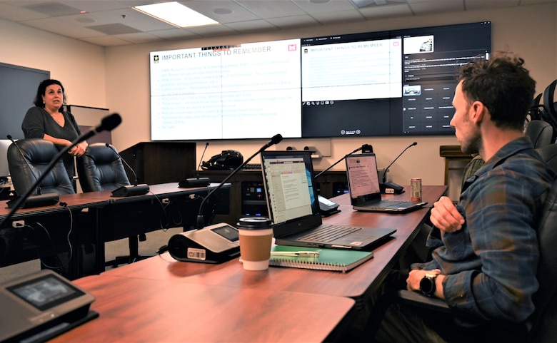 A white woman stands to face a sitting man across a set of tables with computers on them.