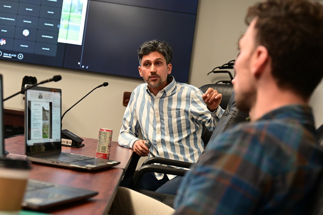 Two white men face each over across a wooden tables with computers on them.