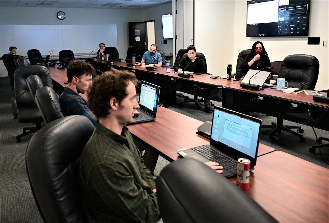 Eight white men and women sit around wooden tables with computers in front of them and TVs on the wall behind them.