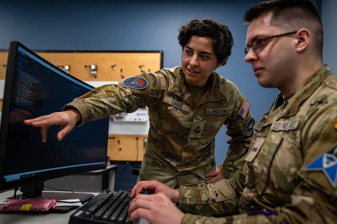 A guardian stands and points to a monitor as a service member sitting by it observes.