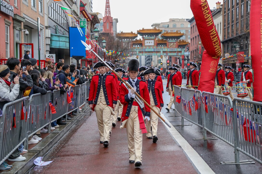 Soldiers in colonial uniforms march in a parade on a city street as spectators watch from behind barricades.