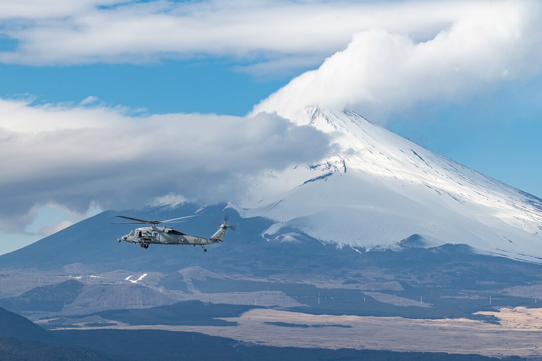 A military aircraft flies in front of a mountain during daylight with a low cloud above.