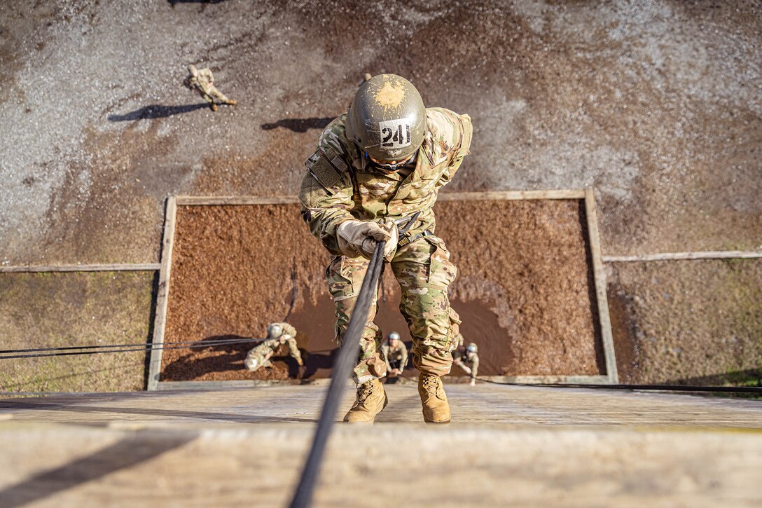 A uniformed service member rappels down a tower during daylight as others on the ground watch.
