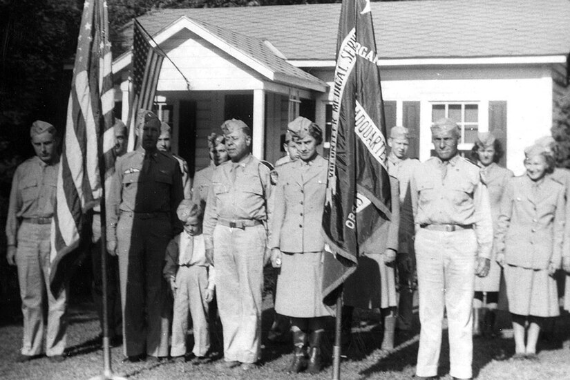 A group of people in uniforms stand at attention behind two flags.