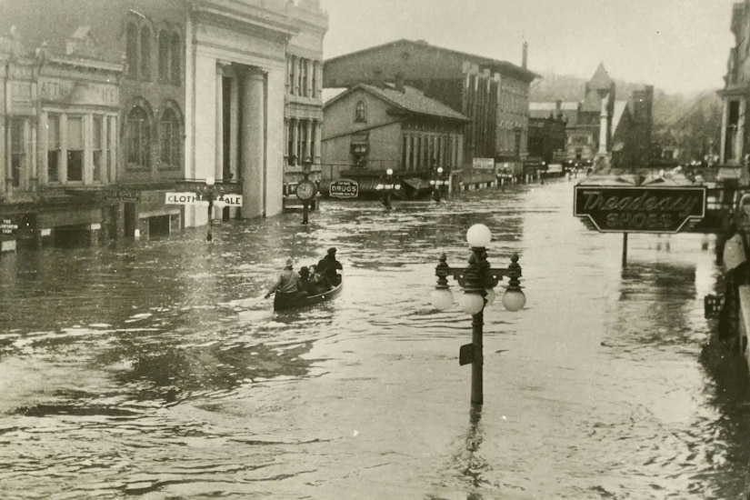 A street in a town is flooded with water. A canoe rows through it.