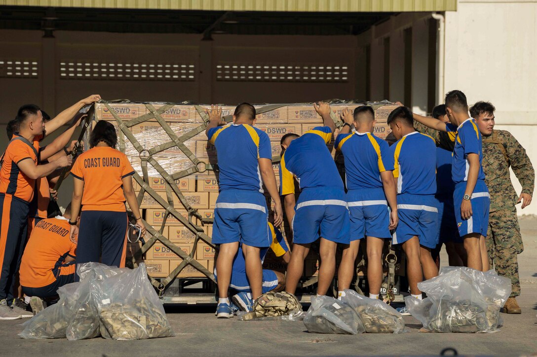 U.S. Marines and Philippine service members hold onto a pallet filled with boxes with military uniforms in clear bags behind them.