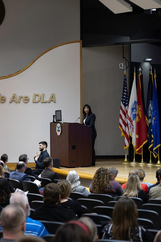 A dark skinned, dark haired woman in a black suit delivers an address on stage in an auditorium. There are several flags behind her. She speaks to an audience seated in the red fabric chairs of the auditorium. In front of her is a light skinned man who is a Deaf interpreter in a black shirt and pants.
