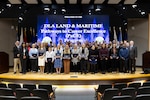 A large group of people are gathered on stage in a large auditorium. They stand in front of a group of flags on stage. The stage is of a yellow wood and several rows of seats are shown. They all hold certificates of graduation except for a few. They are men and women of all ages and are wearing business attire.