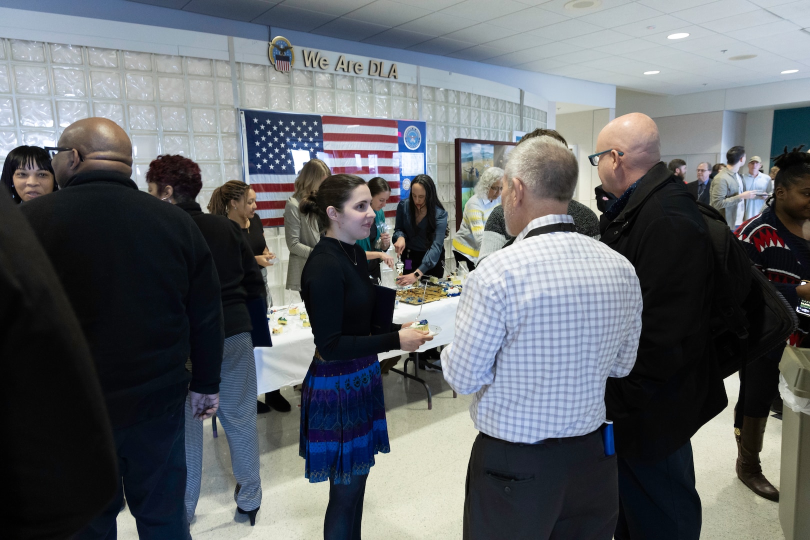 A large group of people are gathered in a open office area with marble floors and glass bubble walls. There is a table up against the bubble glass wall with cake and punch. A focus of the photo is a dark haired woman with her hair in a bun wearing a black shirt and blue skirt talking to two light skinned men in office attire (dress shirts and dress pants). She holds her graduation certificate.