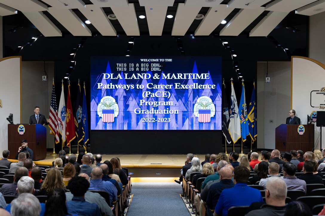 A large group of people are seated in a auditorium attending a graduation ceremony. Photo is shot from the back looking at the audience from behind to the stage in the distance.