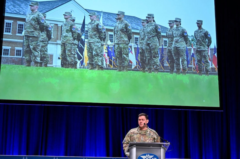 A man in uniform stands at a lectern. Troops stand in formation in the picture on the screen behind him.