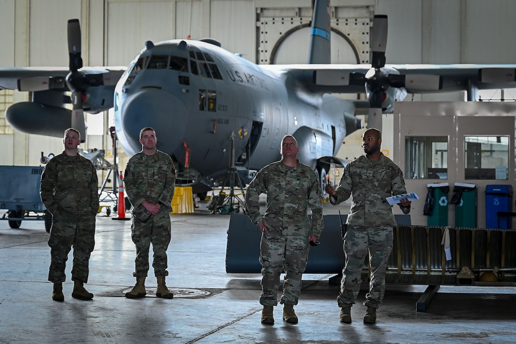 Four Airmen stand in a hanger with a C-130H Hercules aircraft behind them.