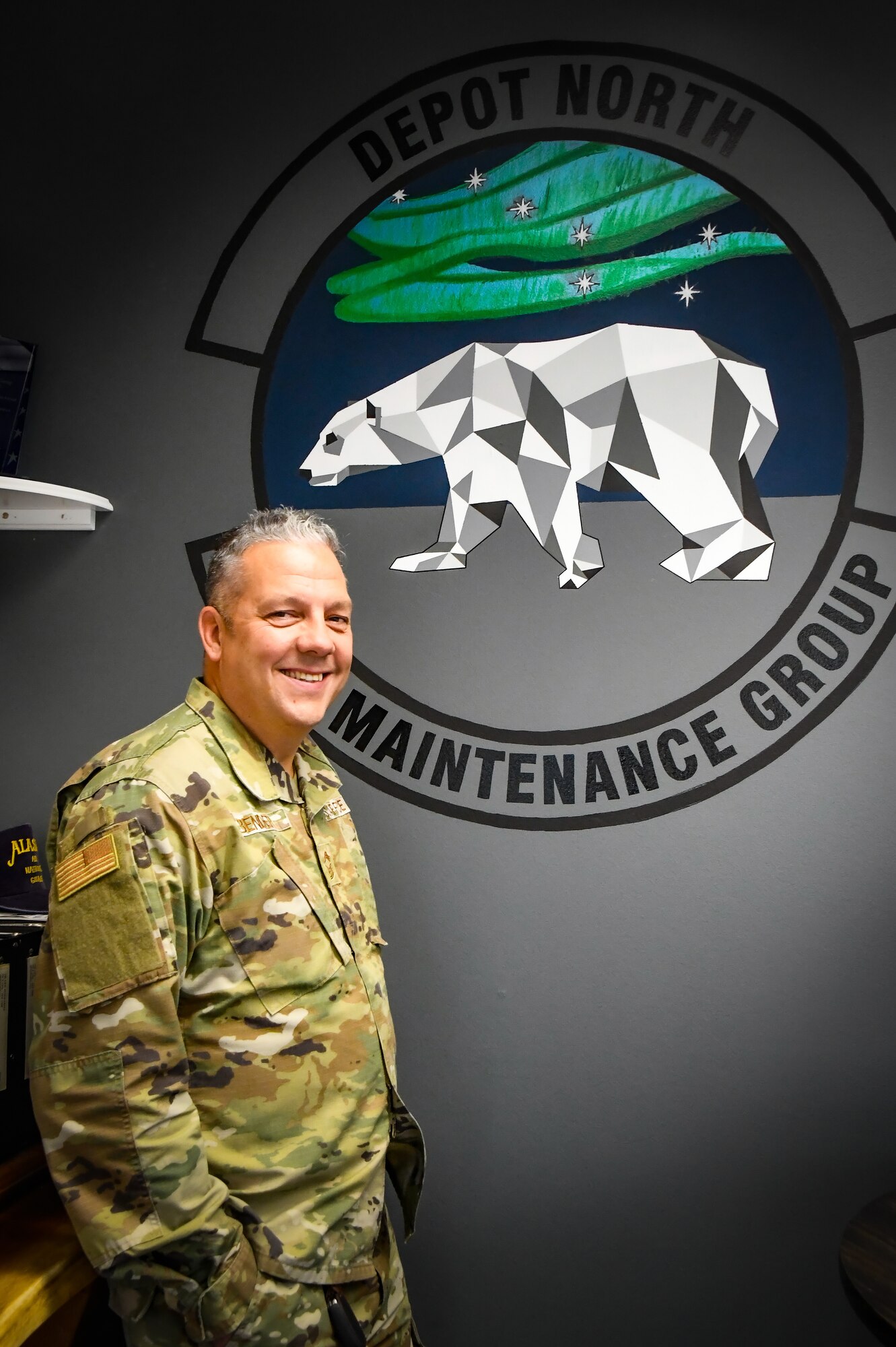 U.S. Air Force Chief Master Sgt. George Bender of the 168th Wing, 168th Maintenance Group, stands in front of his art mural representing the MXG patch, hanging in the MXG hangar at Eielson Air Force Base.