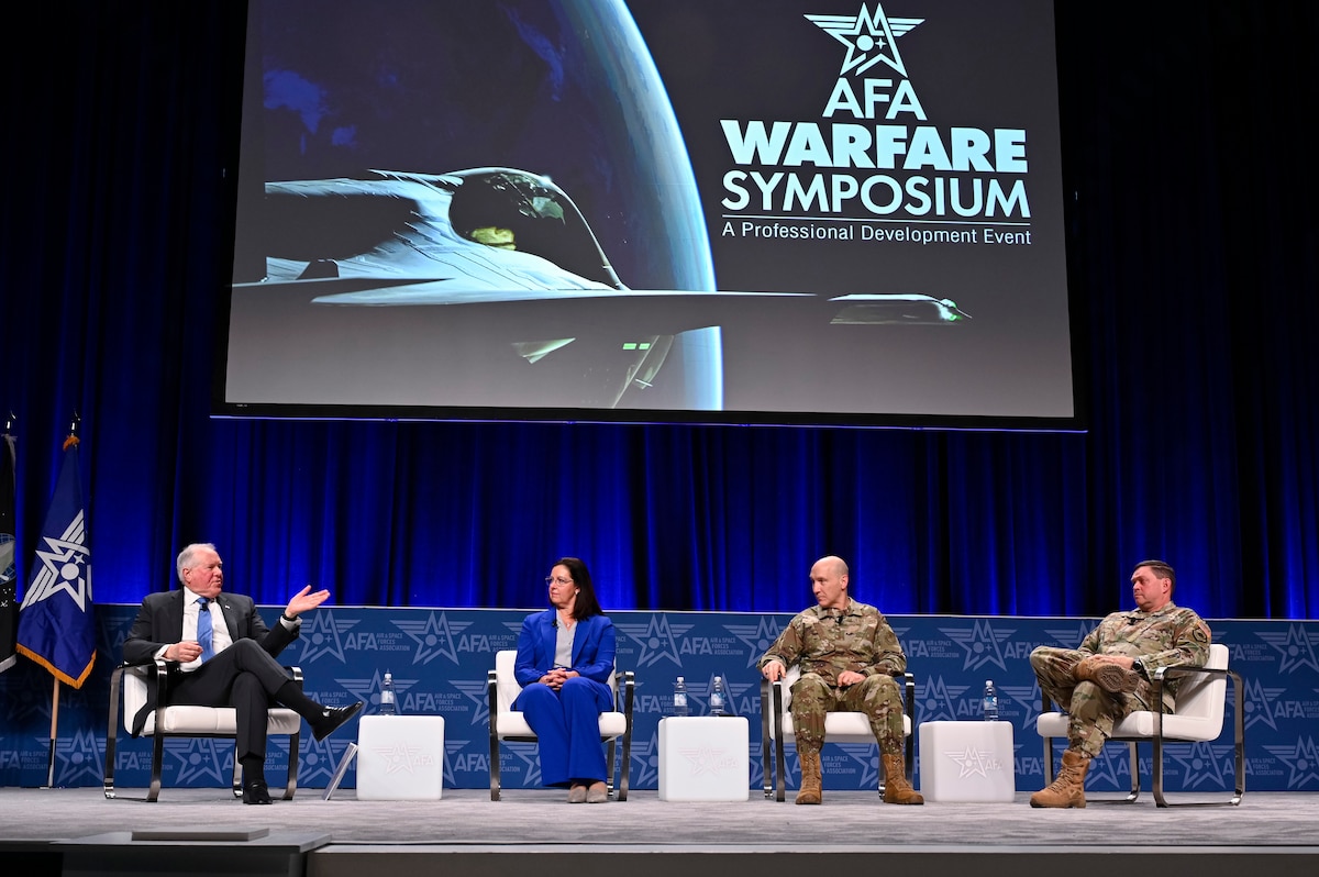 Four people, two in uniforms, sit in chairs on a stage. Behind them a projection screen has the words "AFA Warfare Symposium -- A Professional Development Event."