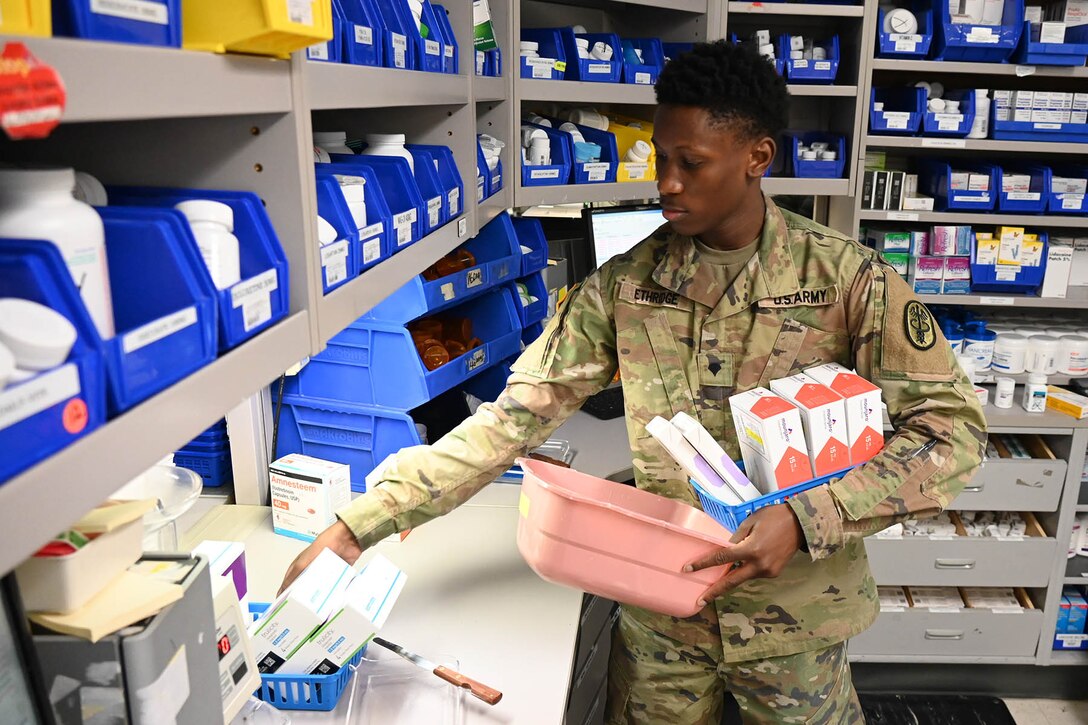 A soldier holds boxes of medication in a space filled with shelves of colored bins holding medications.