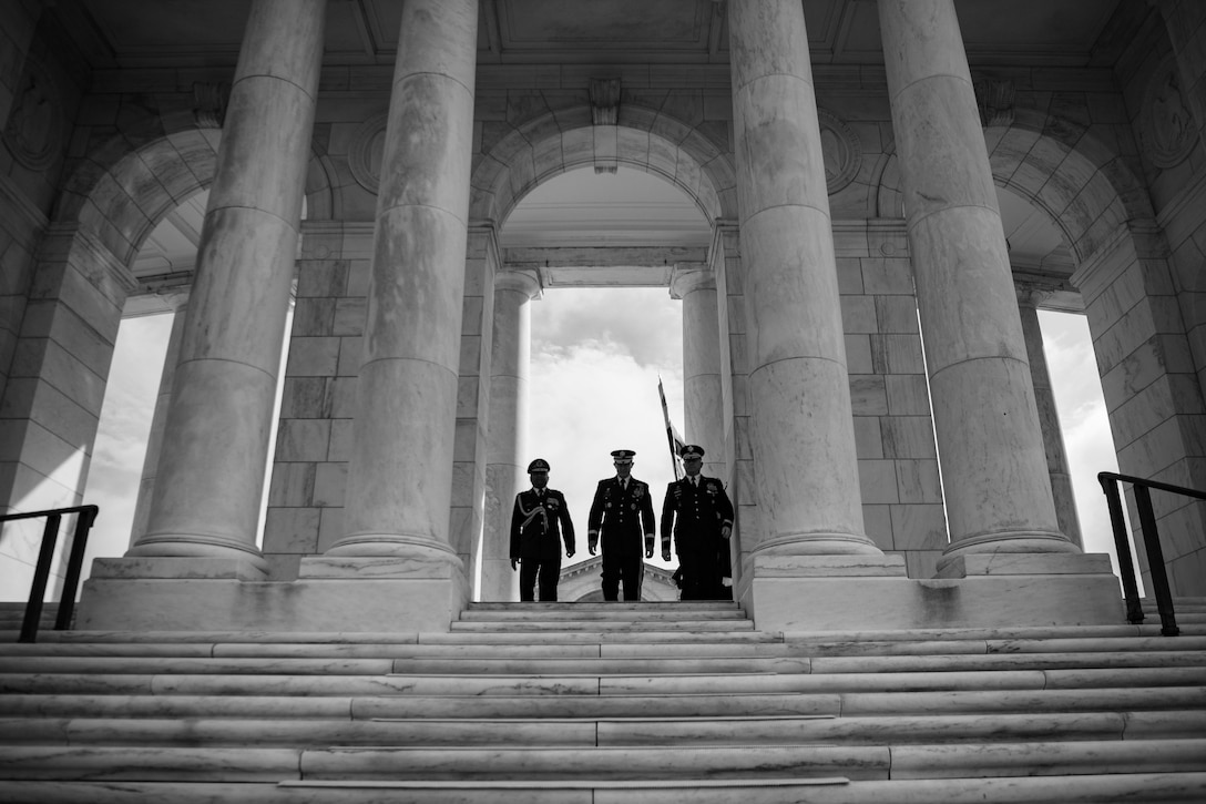 Three military officers stand at the top of a the staircase to a memorial building with two large columns on each side.