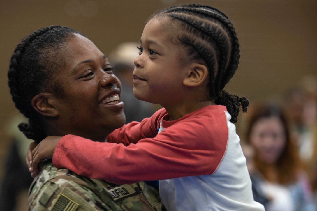 A soldier smiles while holding a child whose arms wrap around her shoulders.