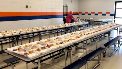 Rows of filled food trays are laid out on a table in what appear to be a school cafeteria.