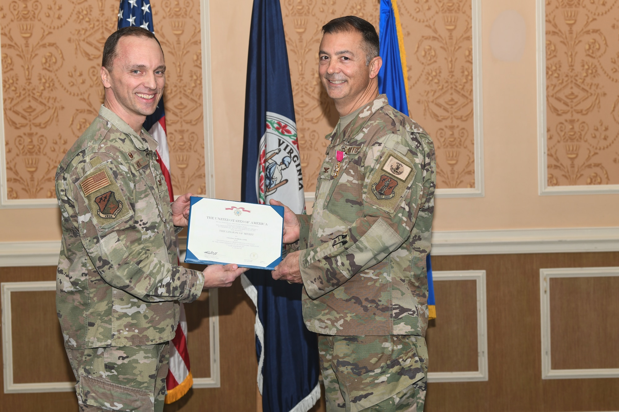 Two military members holding a certificate for the Legion of Merit, smiling for photo.
