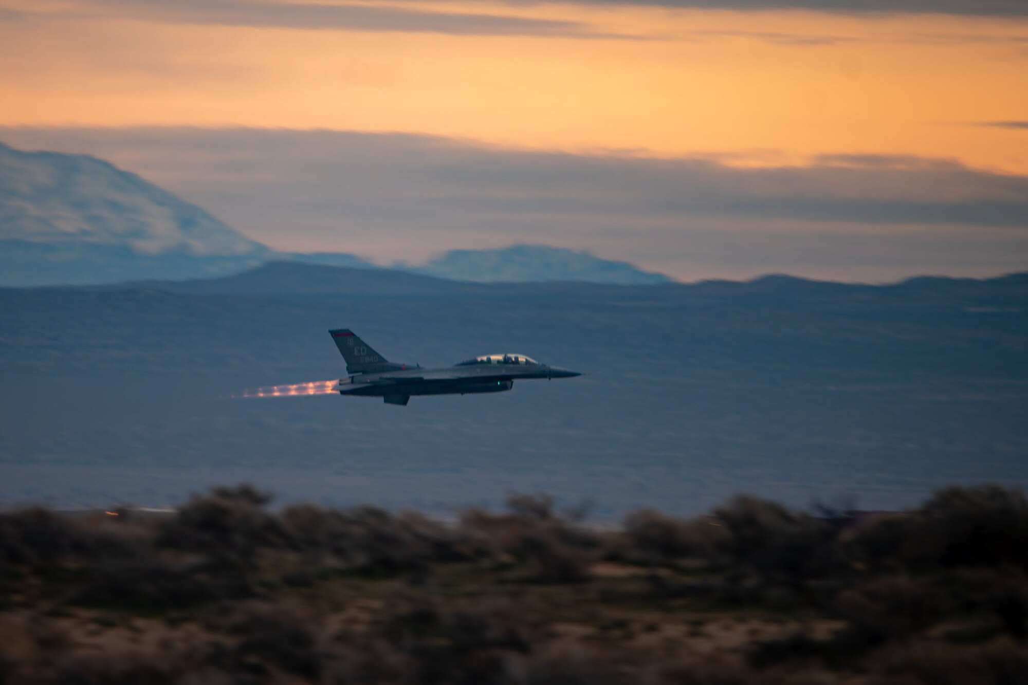 F-16 aircraft flies right with sunset and mountains in the background