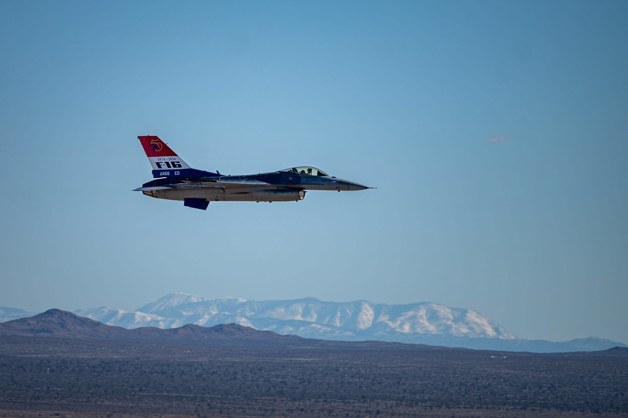 F-16 with red white and blue tail flies to the right with mountains in the background