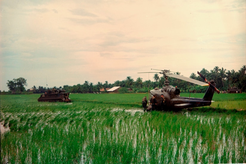 A helicopter and a military vehicle sit in tall grass in a field.