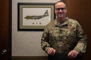 A man smiles, posing for his picture. He is dressed in a military uniform that reads "Borden" and "U.S. Air Force" with different patches visible within the picture. He is wearing plastic, black-framed glasses. In the background, a digital rendering of a plane, contained in a wooden frame.