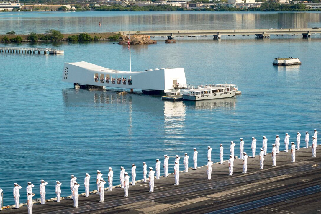 Dozens of sailors lining the edge of the deck of a ship salute the floating USS Arizona memorial.