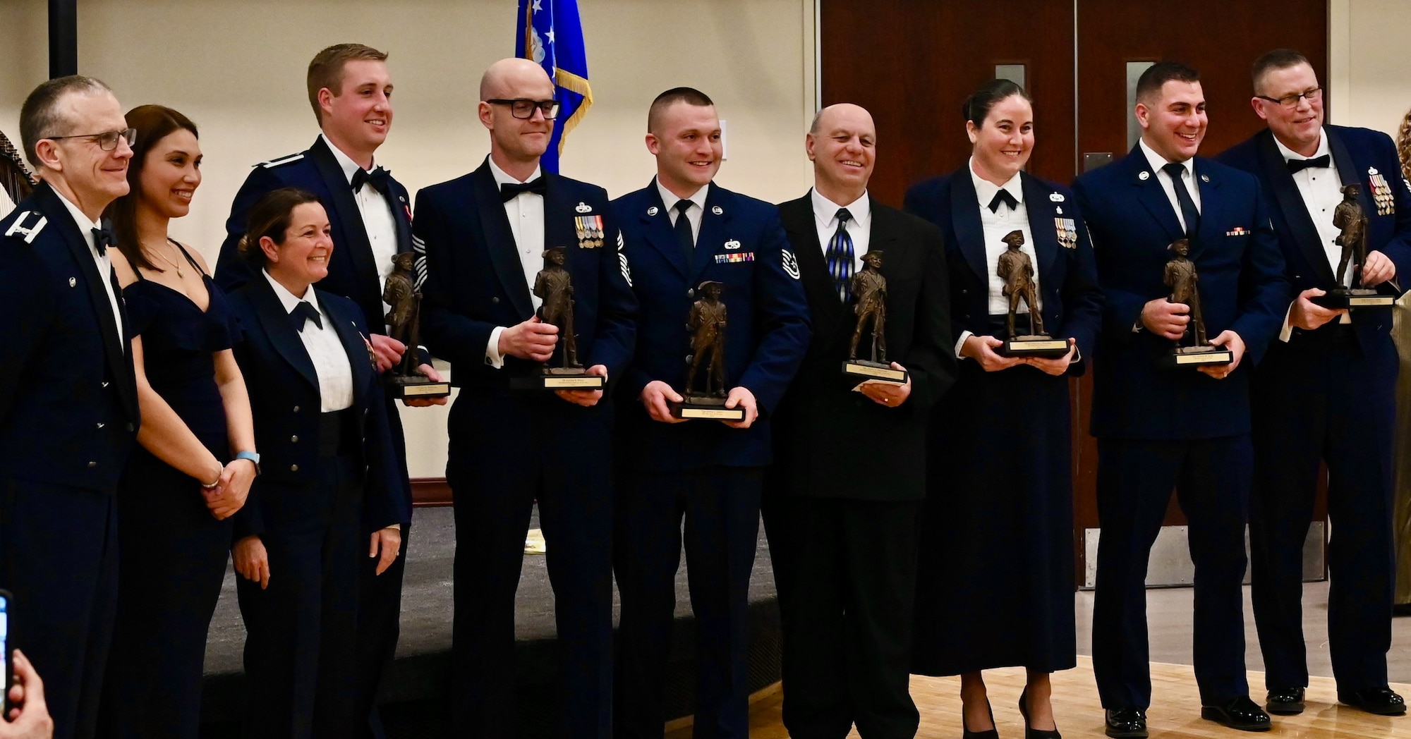 Group of people in military tuxes and formal evening wear pose for a photo, with some of them holding trophies.