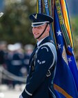 A member of the U.S. Air Force is dressed in dark blue ceremonial service dress and is smiling while holding the blue U.S. Air Force flag