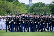 Army Soldiers are marching in rows across a green lawn. They are all wearing service dress uniforms and have a rifle on their right shoulder.