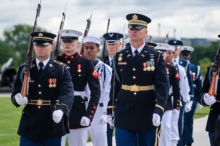 Members from various branches of the military are marching toward the camera. Most have a rifle on their right shoulder.