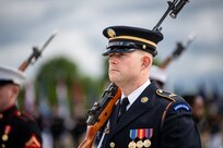 Soldier wearing black dress uniform and hat has rifle on right shoulder.