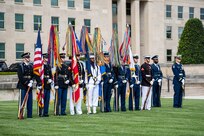 Service members from each branch of the military are standing at parade rest while holding the flags from each branch in front of the Pentagon. On each end is one service member holding a rifle, and there are two service members on the far right who are holding swords.