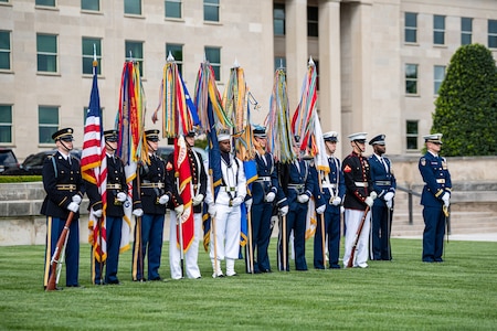 Service members from each branch of the military are standing at parade rest while holding the flags from each branch in front of the Pentagon. On each end is one service member holding a rifle, and there are two service members on the far right who are holding swords.
