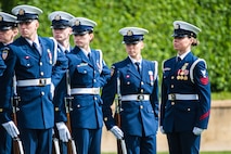 Members of the Coast Guard are in service dress uniforms. They are all looking off the left as one member looks on. Each has a rifle at their side.