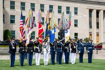 Service members wearing dress uniforms from each branch of the military are carrying each branches flag while standing in a straight line in front of the Pentagon. On each end of the line is a service member carrying a rifle, and two other service members on the far right are carrying swords.