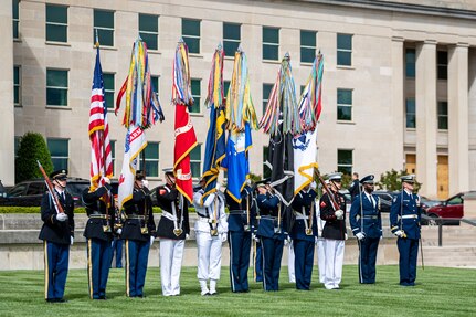 Service members wearing dress uniforms from each branch of the military are carrying each branches flag while standing in a straight line in front of the Pentagon. On each end of the line is a service member carrying a rifle, and two other service members on the far right are carrying swords.