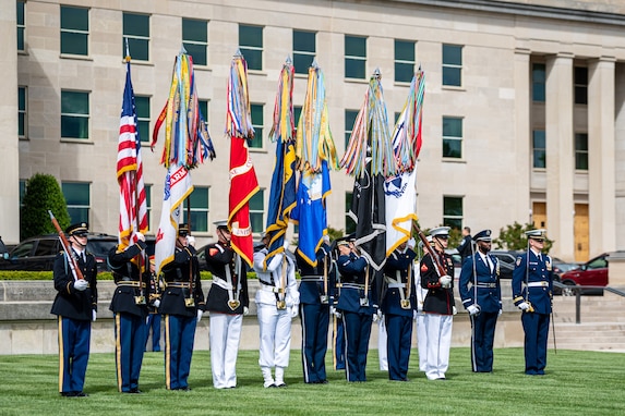 Service members wearing uniforms from each branch are carrying each flag while standing in front of the Pentagon. On each end of the line is a service member carrying a rifle, and two other service members on the far right are carrying swords.