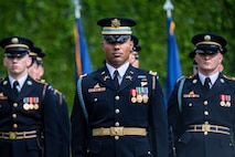 Soldiers in dark blue Army dress uniforms and hats are standing at attention during a ceremony. Behind them are some flags and a large green hedge.
